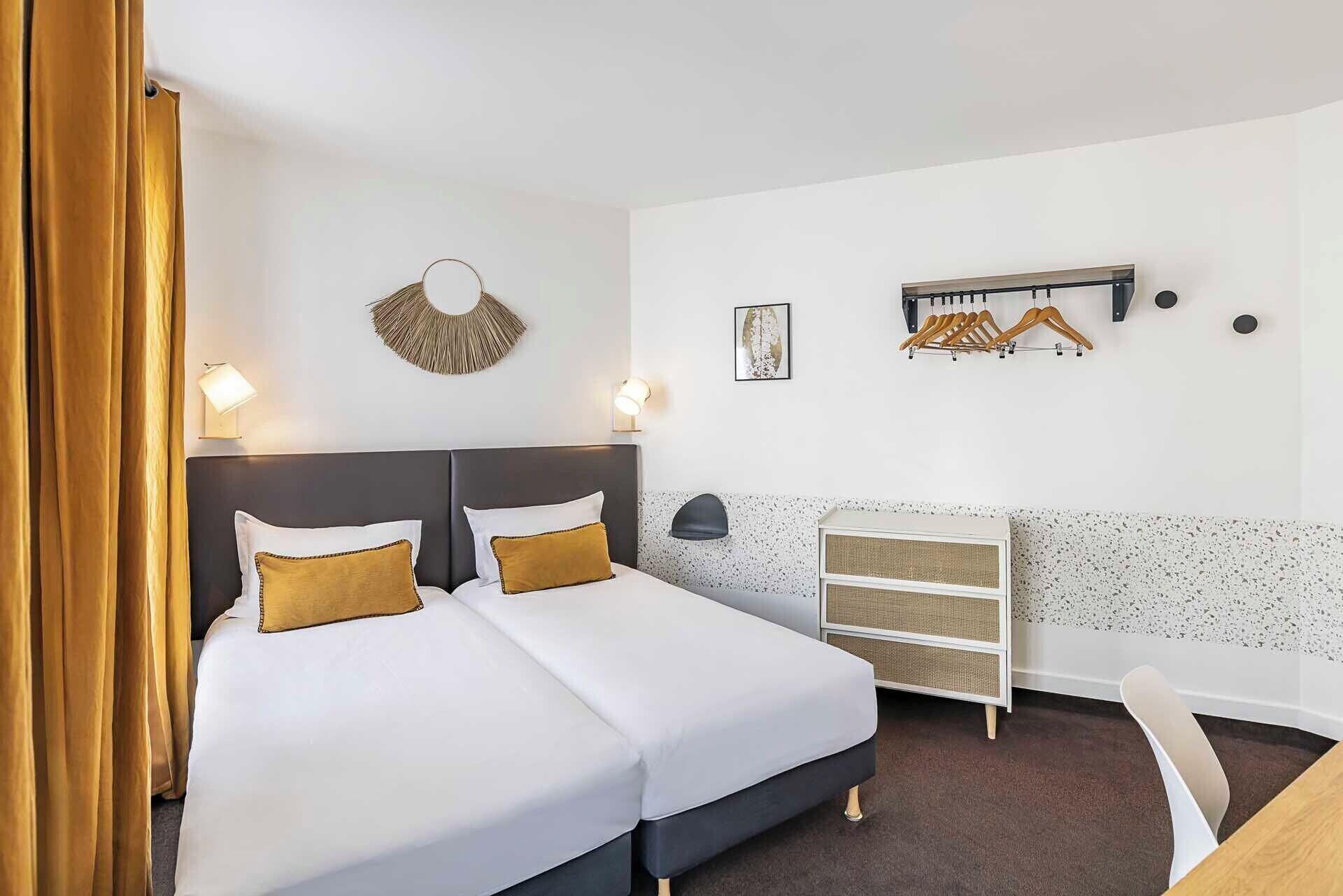 A hotel room with a double bed, a desk, a chair, a wall-mounted coat rack, and a small shelf.