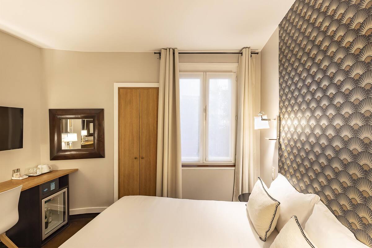 A hotel room with a bed, a wooden desk with a mirror, a minibar, and a window with curtains.