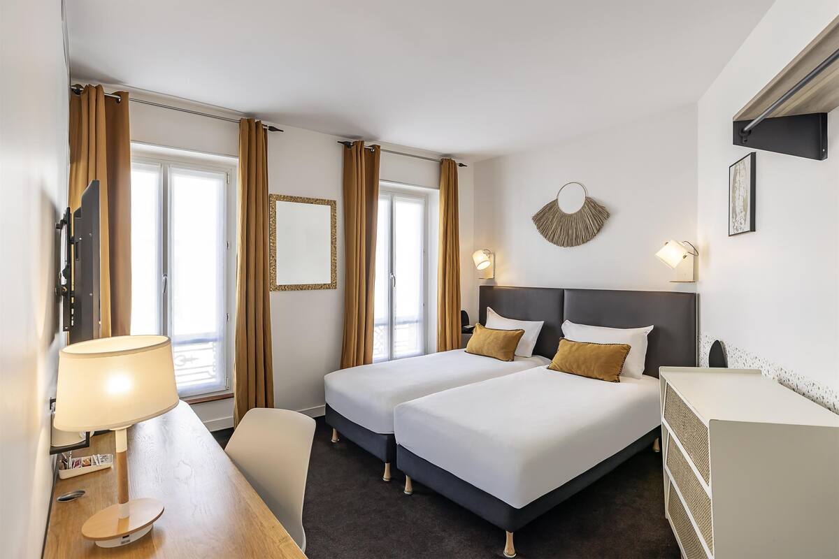 A hotel room with two beds, a desk with a lamp, a chair, a TV, and a window with curtains.