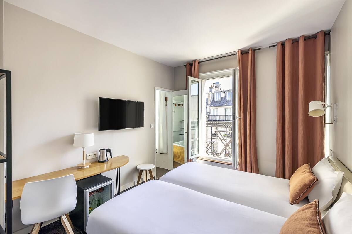 A hotel room with two beds, a desk with a lamp, a TV, and a view of a balcony with curtains.
