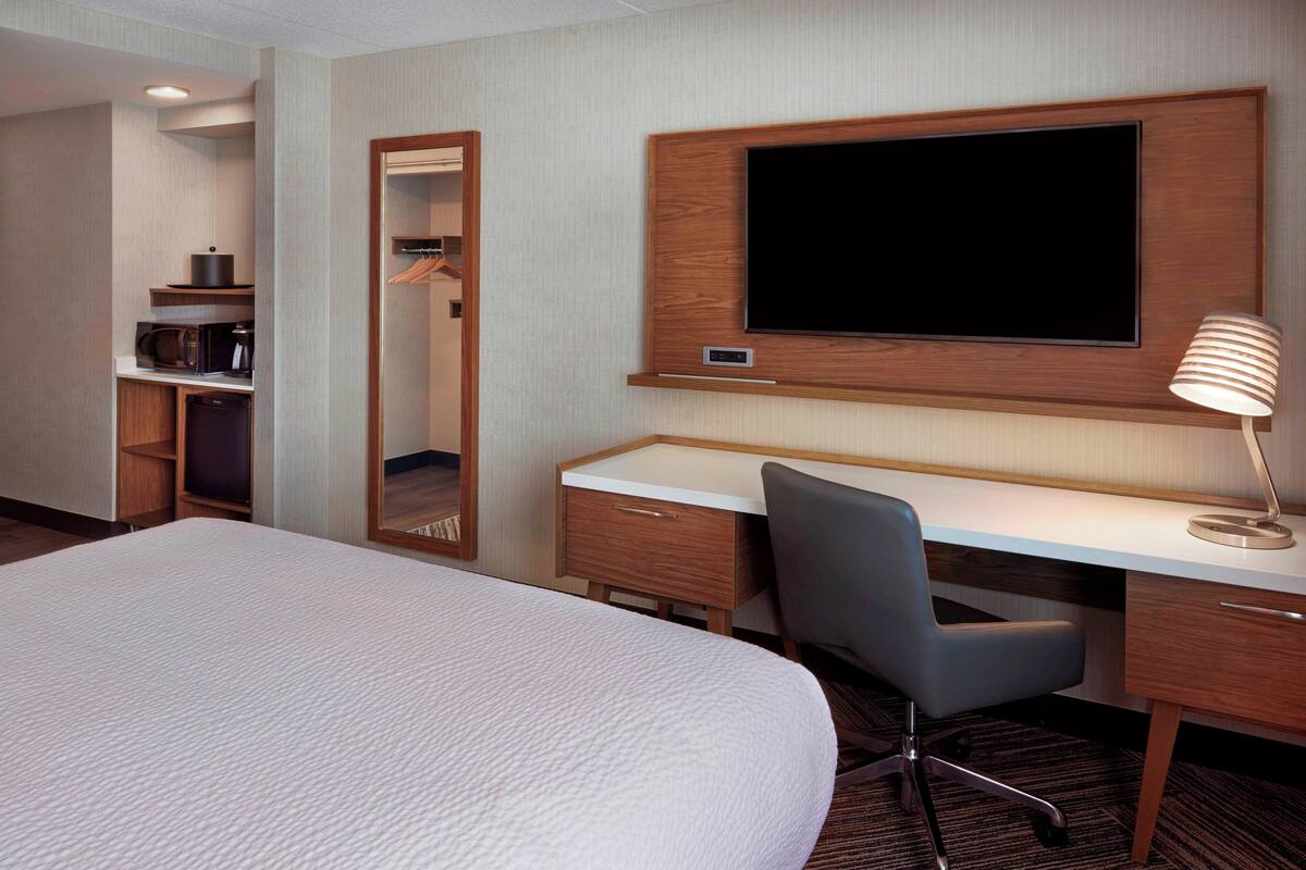 A hotel room with a large bed, a desk with a chair, a flat-screen TV mounted on the wall, a mirror, and a lamp.