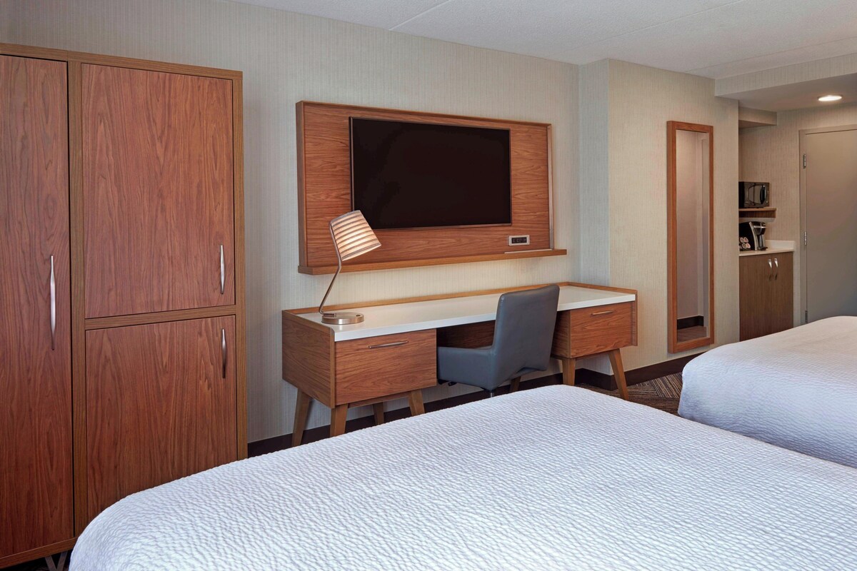 A hotel room with a wooden wardrobe, a desk with a chair, a television, and two beds.
