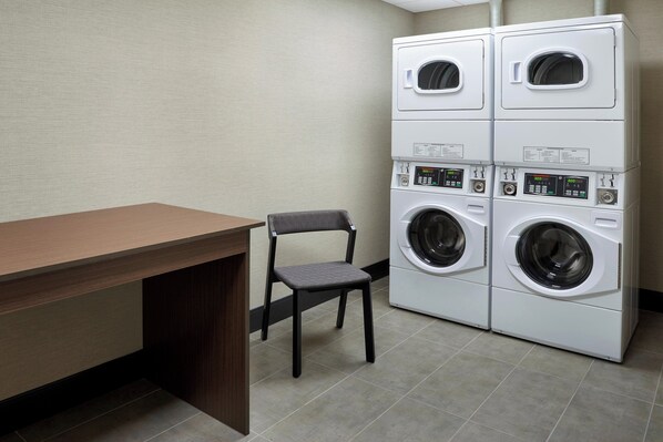 A laundry room with a wooden table and a chair, and a stack of four white front-loading washing machines. - Four Points by Sheraton Hamilton - Stoney Creek (Hamilton)