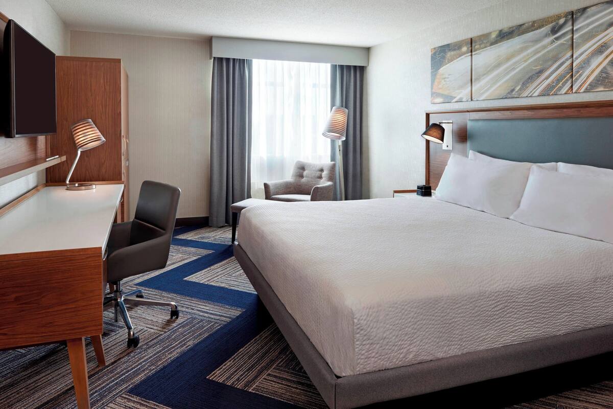 A hotel room with a large bed, a desk, a chair, a TV, and a window with curtains.