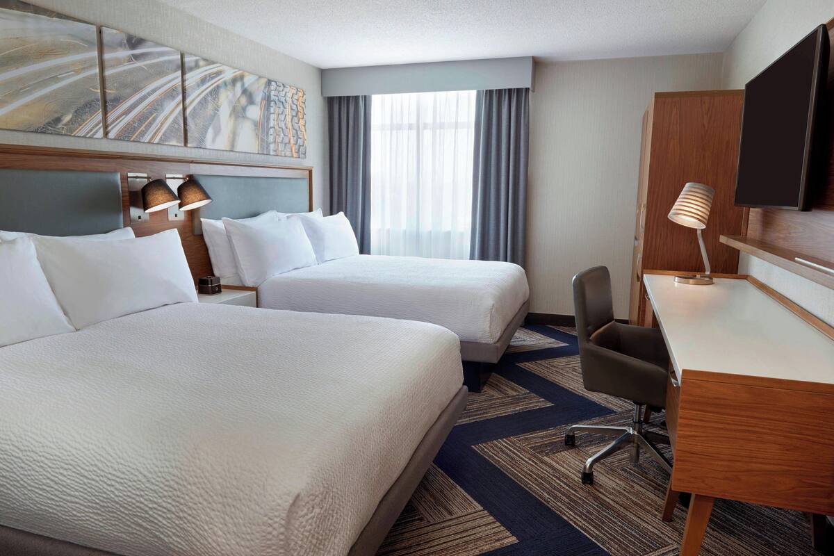 A hotel room with two beds, a desk, a chair, a TV, and a large window with curtains.
