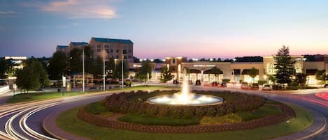 A circular fountain with water jets in the center, surrounded by a landscaped area with trees and shrubs. In the background, there are buildings with signage, including one that reads "New York Company."