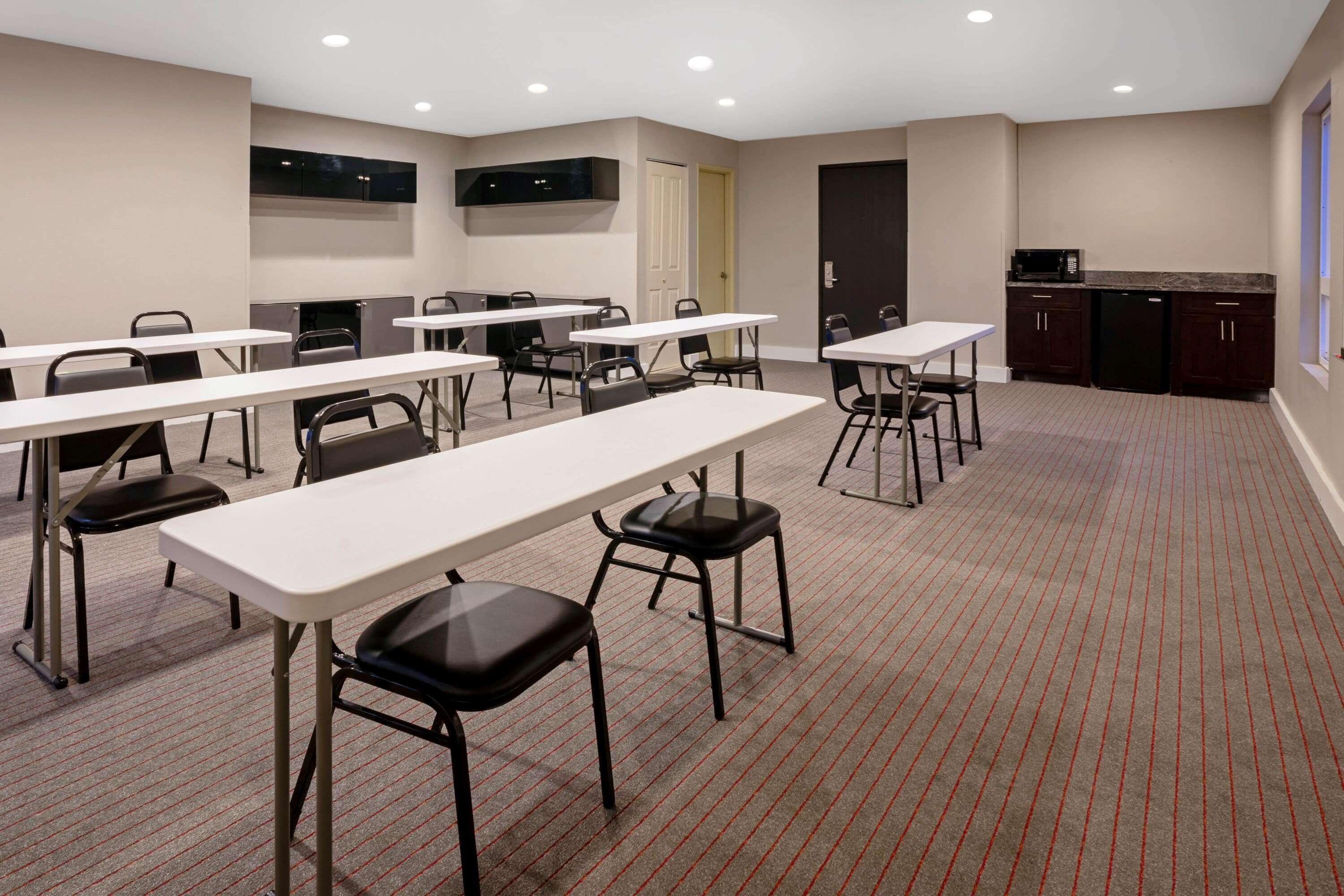 A conference room with multiple tables and chairs, a kitchen area with a microwave and refrigerator, and a carpeted floor.