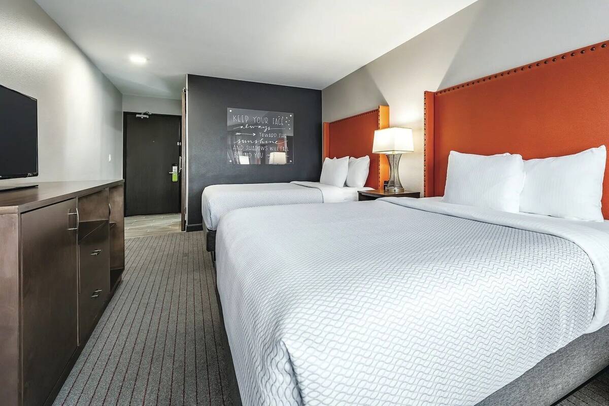 A hotel room with two beds, a desk, a chair, a wall-mounted TV, and a motivational quote on the wall.