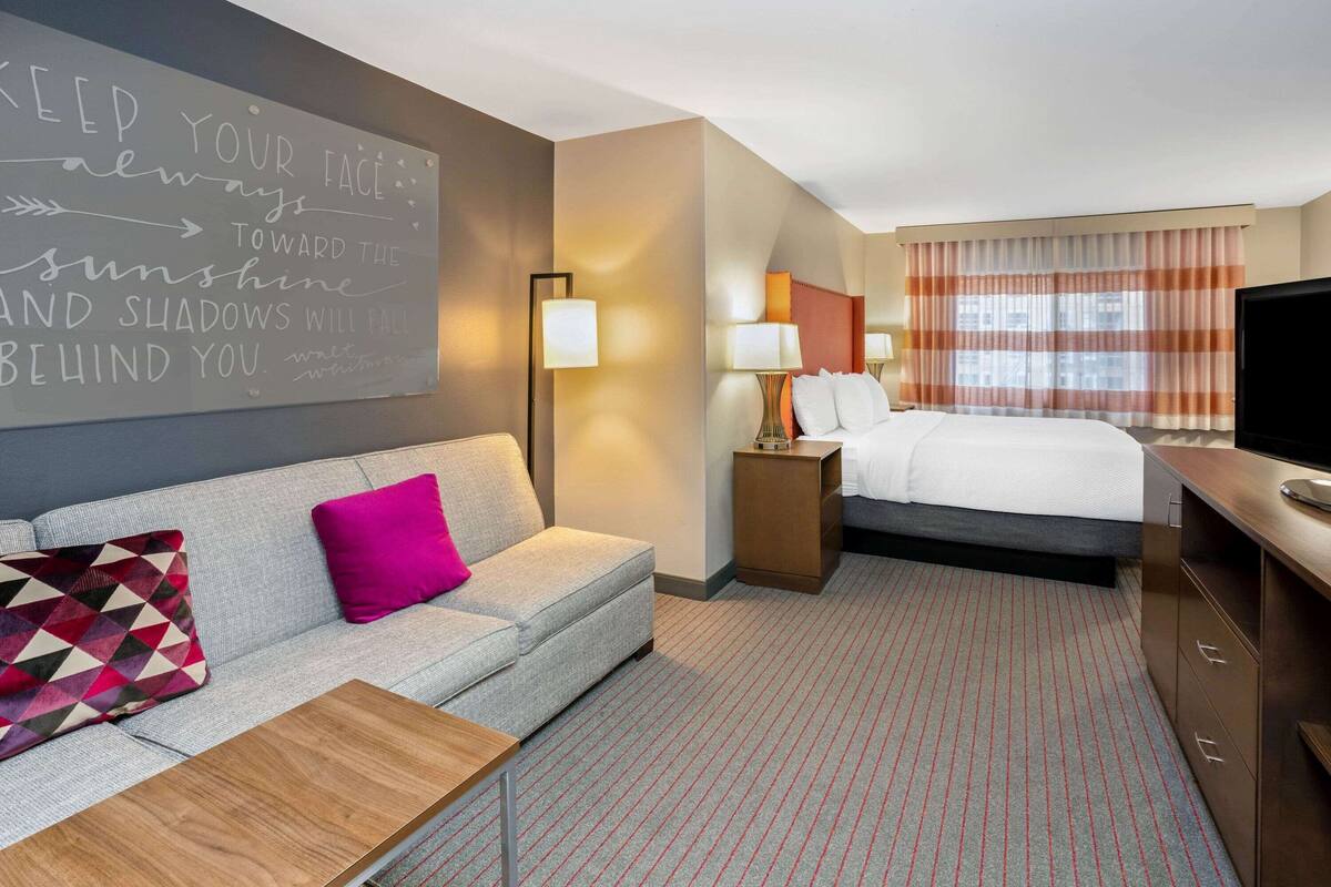A hotel room with a bed, a sofa, a TV, a lamp, and a motivational quote on the wall.