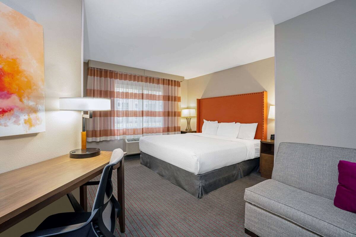 A hotel room with a bed, a desk, a chair, a sofa, and a large window with curtains.