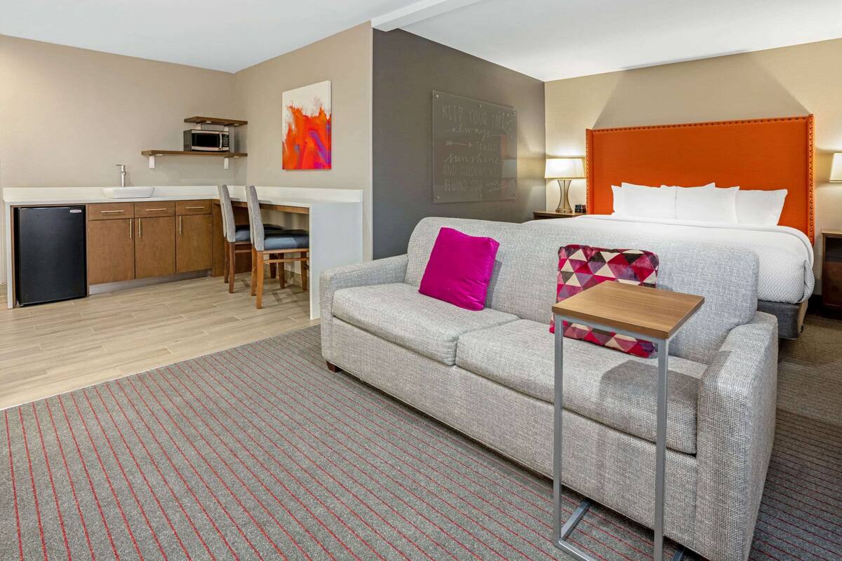 A modern hotel room with a grey sofa, a small wooden table, a bed with an orange headboard, a kitchen area with a microwave, and a red and white patterned pillow on the sofa.
