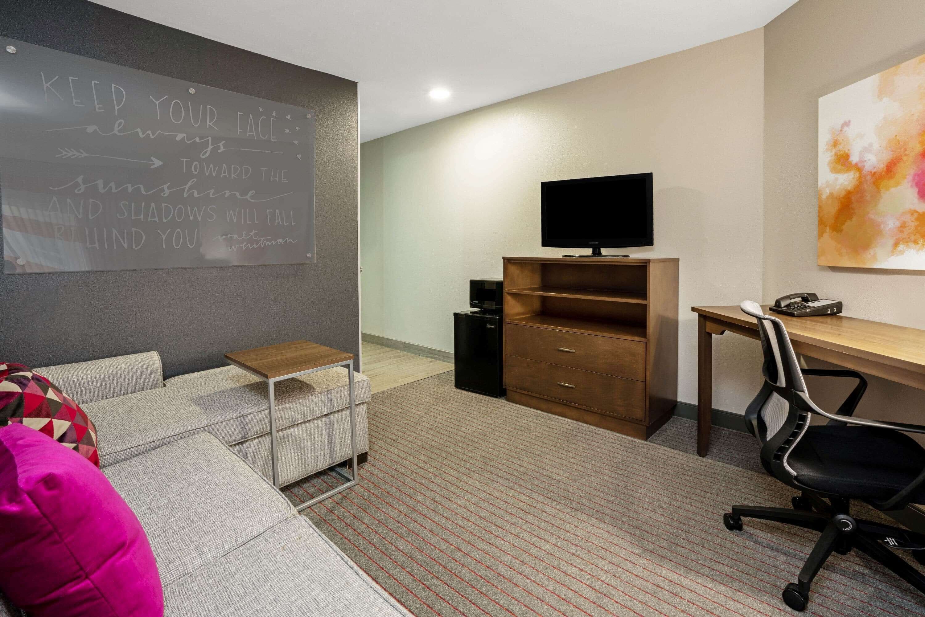 A modern room with a grey couch, a wooden desk, a black office chair, a television, and a chalkboard with motivational writing.