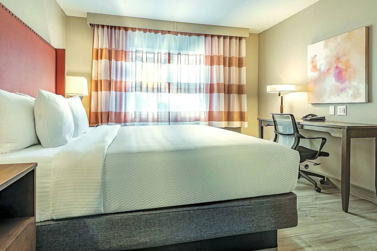 A hotel room with a bed, desk, chair, and a painting on the wall.
