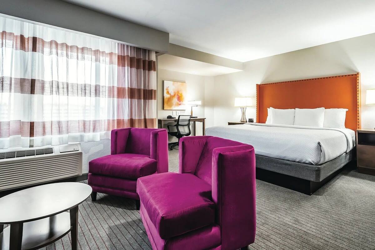 A hotel room with a bed, two purple armchairs, a small table, a desk with a chair, and a large window with curtains.