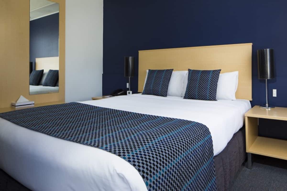 A hotel room with a large bed, a bedside table, a nightstand, a mirror, and a wall-mounted lamp.