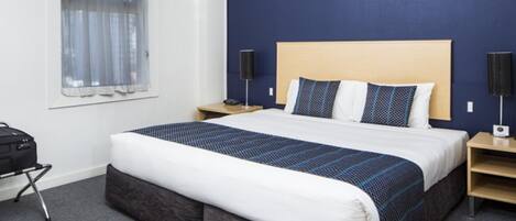 A hotel room with a large bed, a window with curtains, a desk with a lamp, and a suitcase.