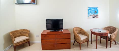 A living room with a TV, a wooden cabinet, wicker chairs, a round table, and a beach-themed poster on the wall.
