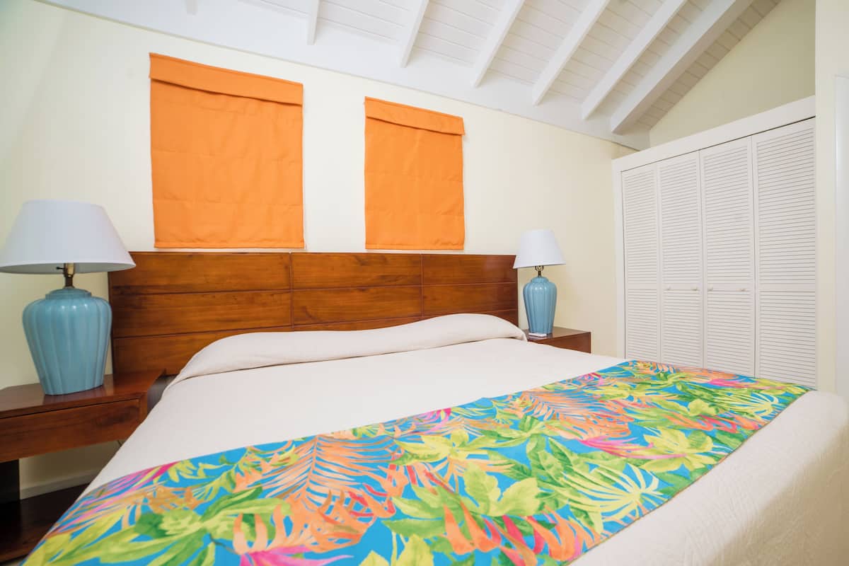 A hotel room with a wooden headboard, two windows with orange blinds, a bed with a colorful floral bedspread, two bedside tables with lamps, and a white closet.