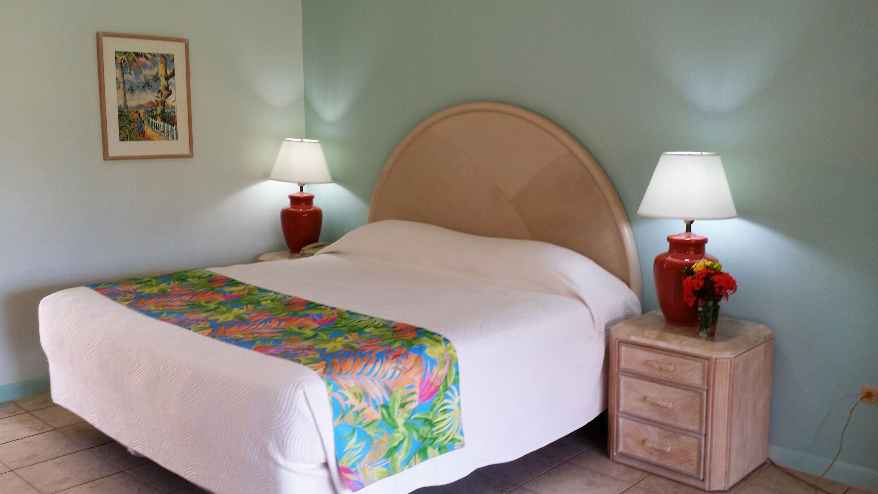 A neatly made bed with a colorful tropical-themed bedspread, two bedside lamps, and a small wooden nightstand with a drawer.
