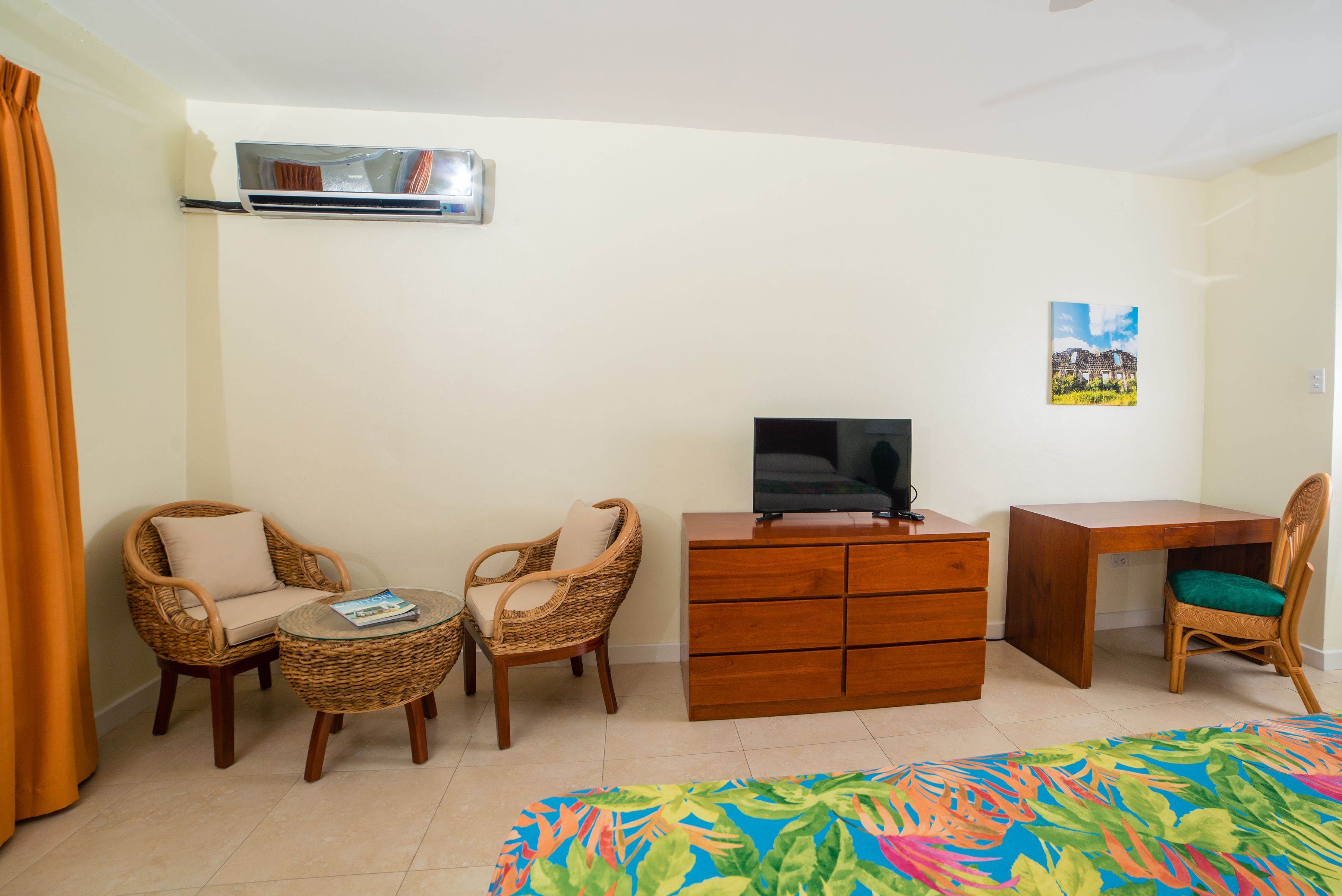 Quarto com cadeiras de vime, uma mesa redonda pequena, cômoda de madeira, televisão e uma cama com colcha colorida.