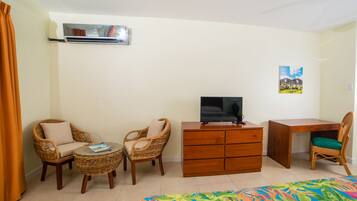 Quarto com cadeiras de vime, uma mesa redonda pequena, cômoda de madeira, televisão e uma cama com colcha colorida.