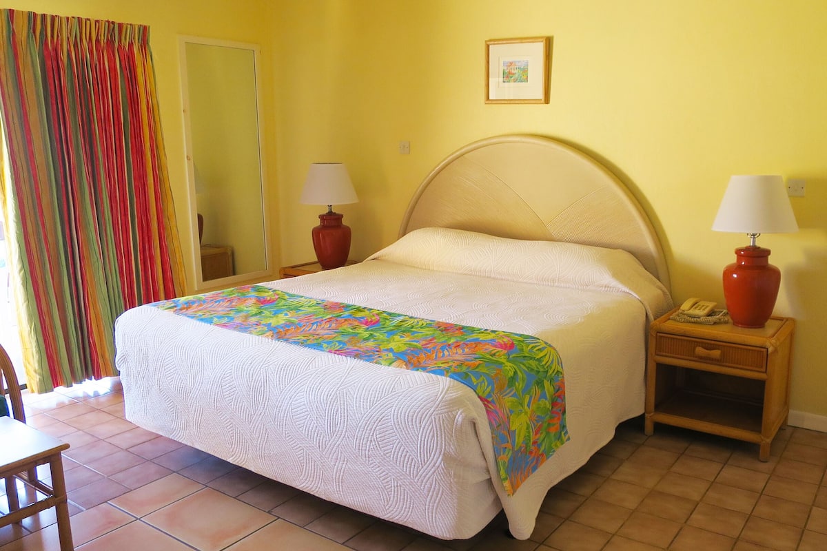 A hotel room with a bed, two bedside lamps, a bedside telephone, a small wooden nightstand, a colorful bedspread, a mirror, and a door with a striped curtain.