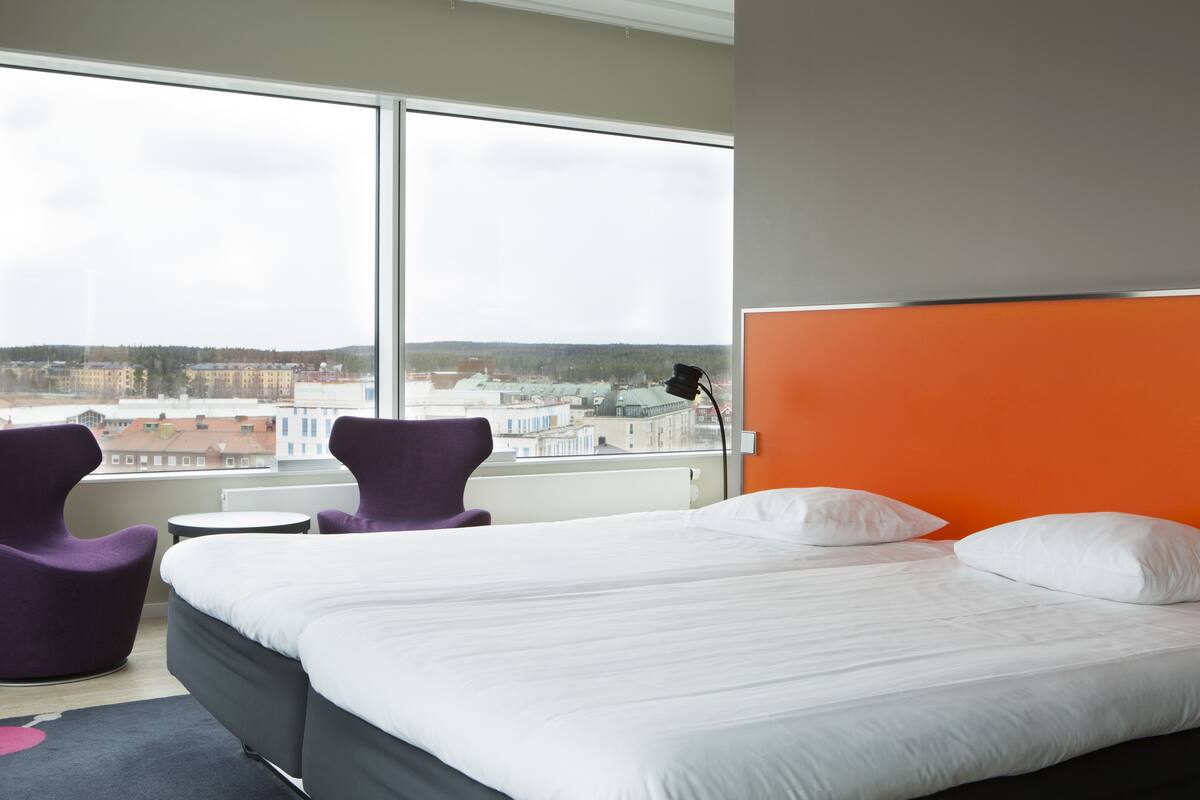 A hotel room with a large bed, two purple chairs, a small table, and a view of buildings through a large window.