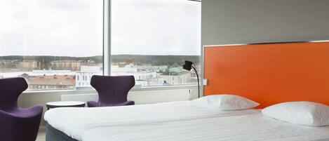 Habitación de hotel con una cama grande, dos sillas moradas, una mesita y vistas a edificios a través de un ventanal amplio.