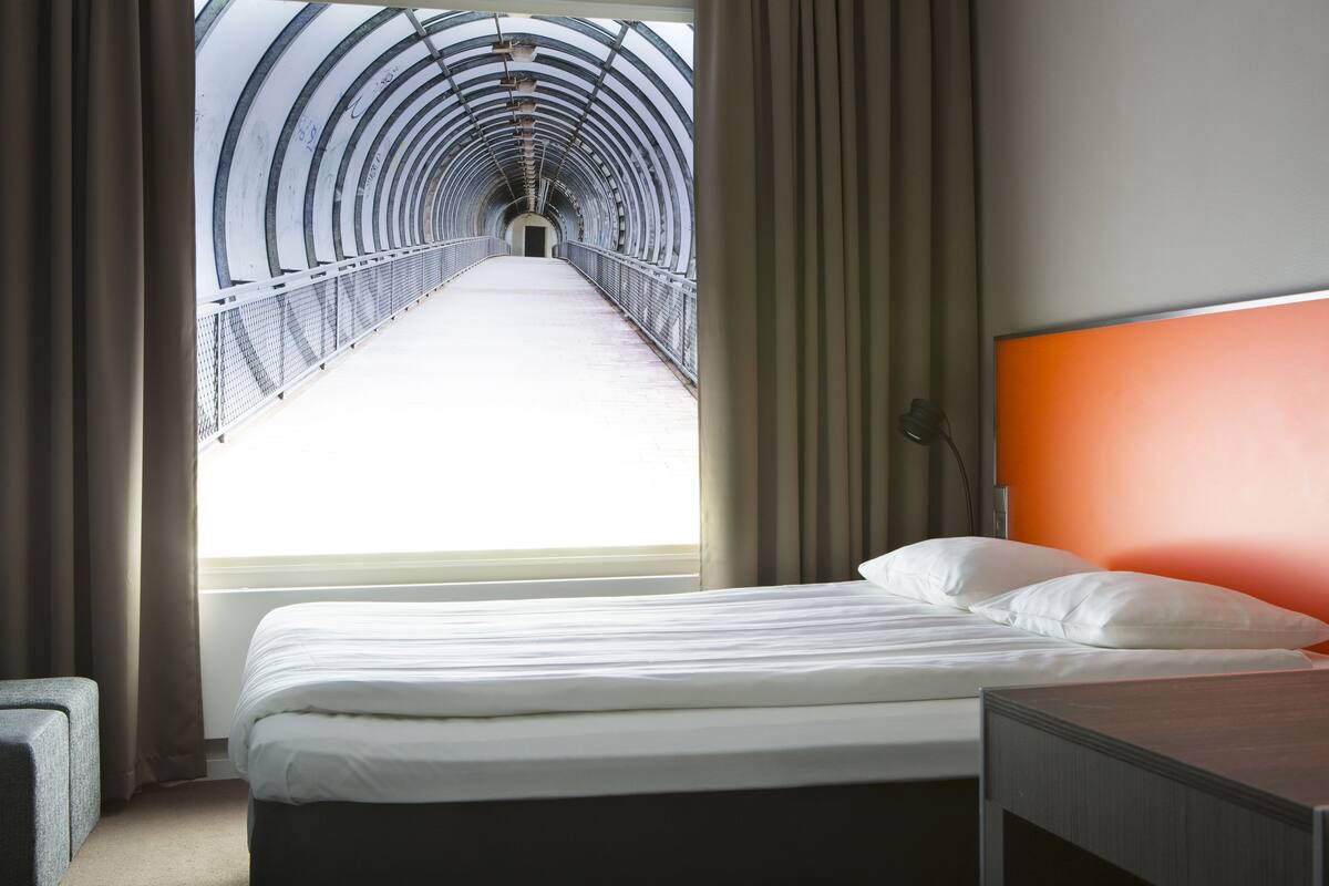 A hotel room with a bed, a nightstand, a window with curtains, and a view of a tunnel.
