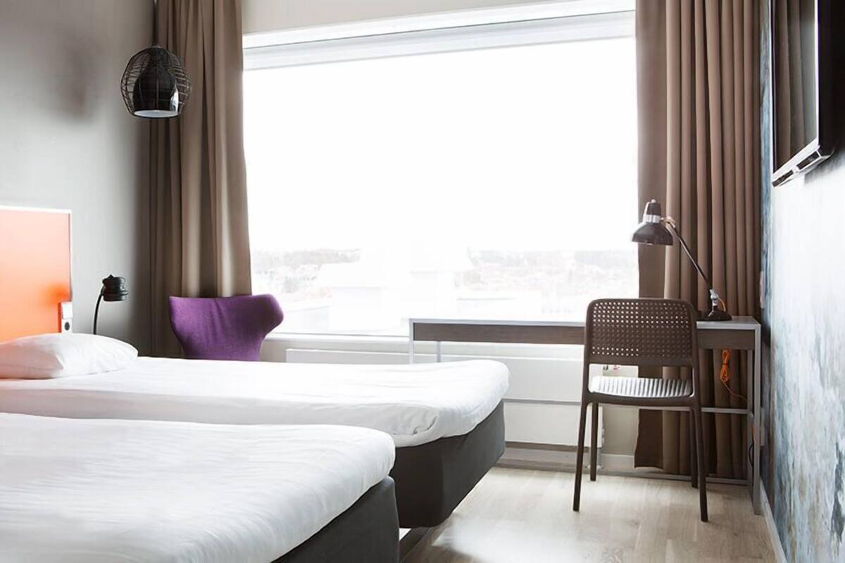 A hotel room with two beds, a desk, a chair, a window with curtains, and a wall-mounted lamp.