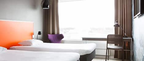 Ein Hotelzimmer mit zwei Betten, einem Schreibtisch, einem Stuhl, einem fenster mit Vorhängen und einer Wandlampe.