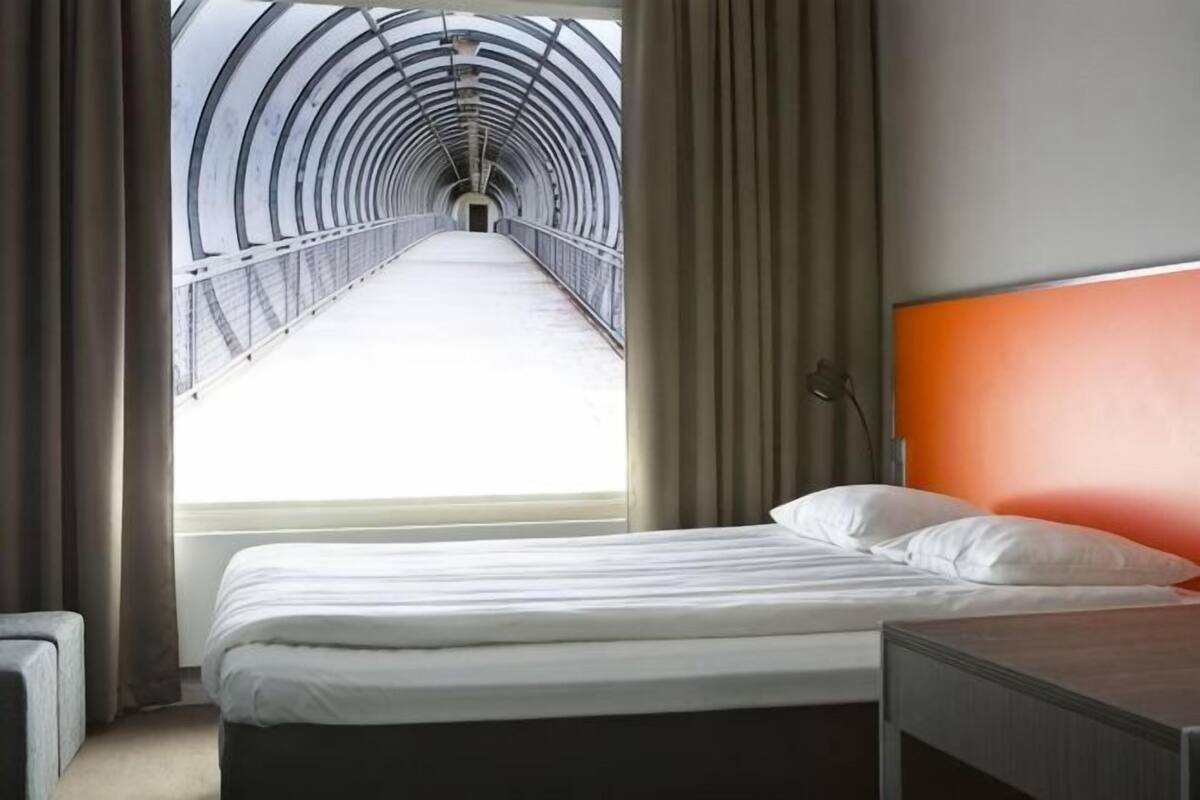 A hotel room with a bed, a nightstand, a lamp, and a view of a tunnel.