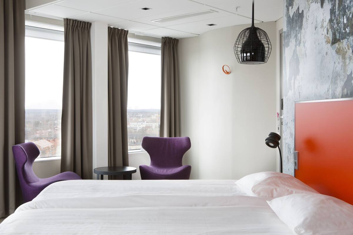 A hotel room with a large bed, two purple chairs, a small table, and a view of the city through large windows.