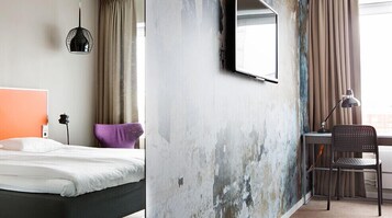 A hotel room with a bed, a desk with a chair, a hanging lamp, and a large mirror on the wall.