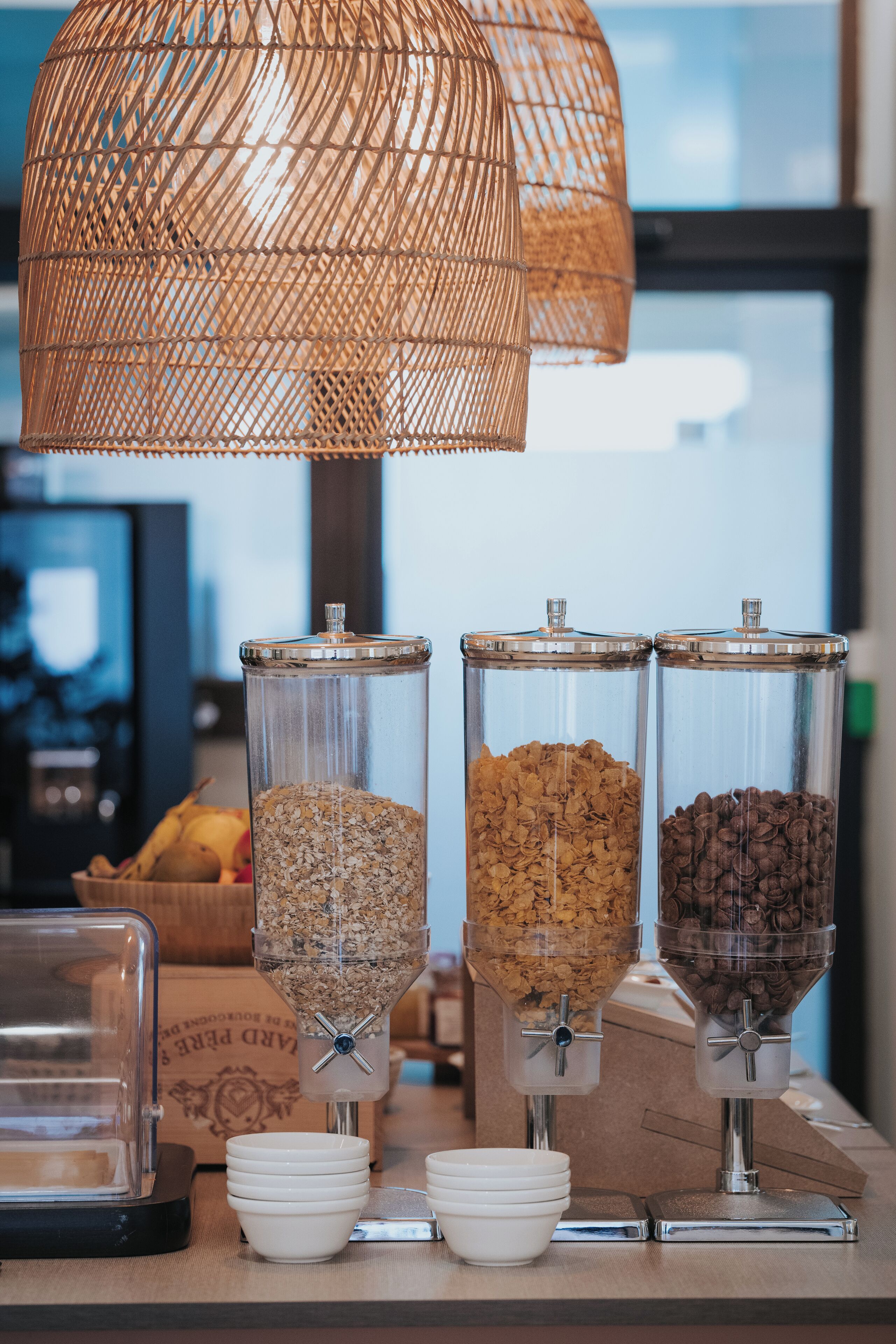 Un comptoir pour le petit-déjeuner avec trois distributeurs de granola et de café, des bols blancs et une lampe en osier.