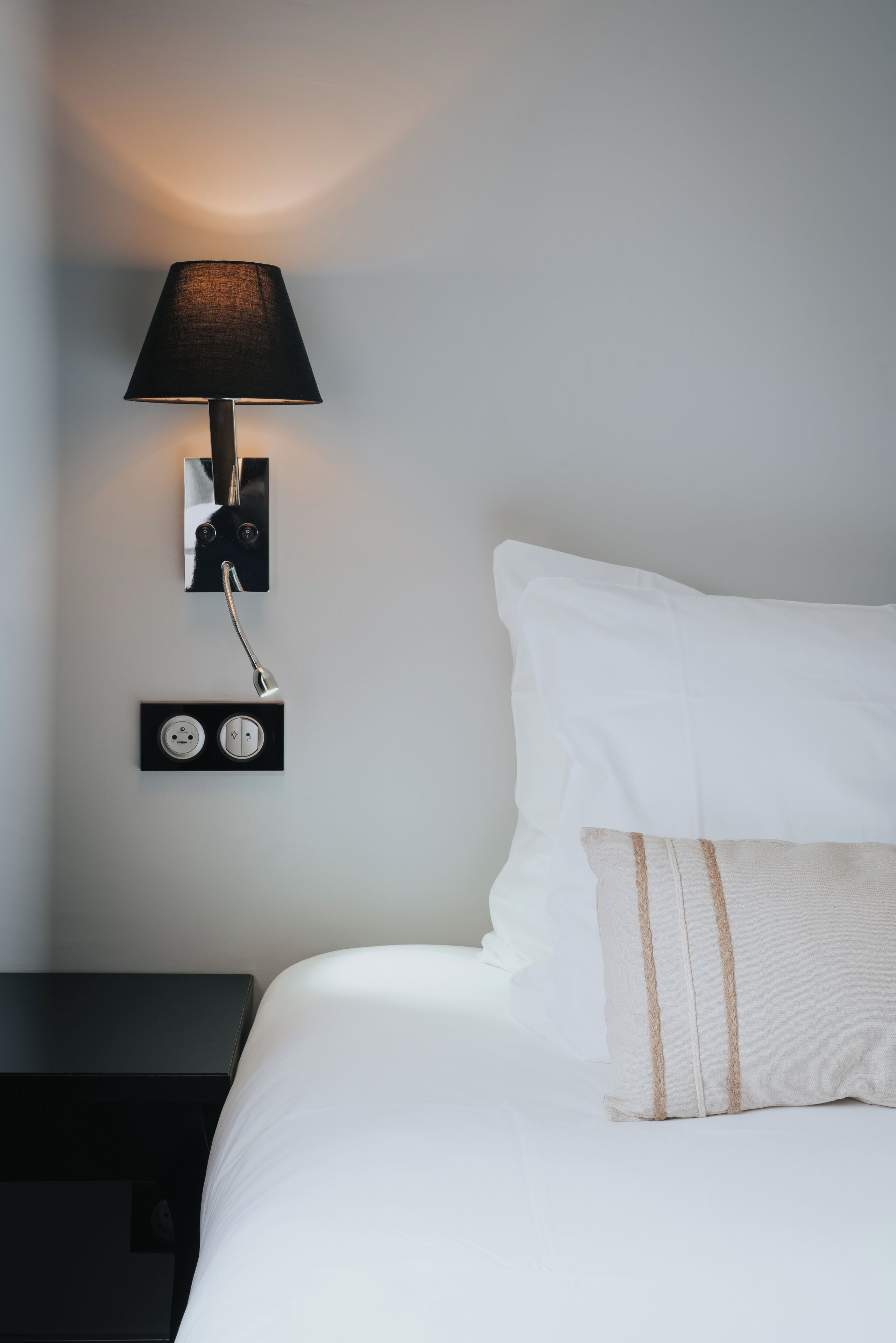Un lit bien fait, avec du linge de lit blanc et un seul coussin décoratif, une table de chevet noire et une lampe murale avec un abat-jour noir.