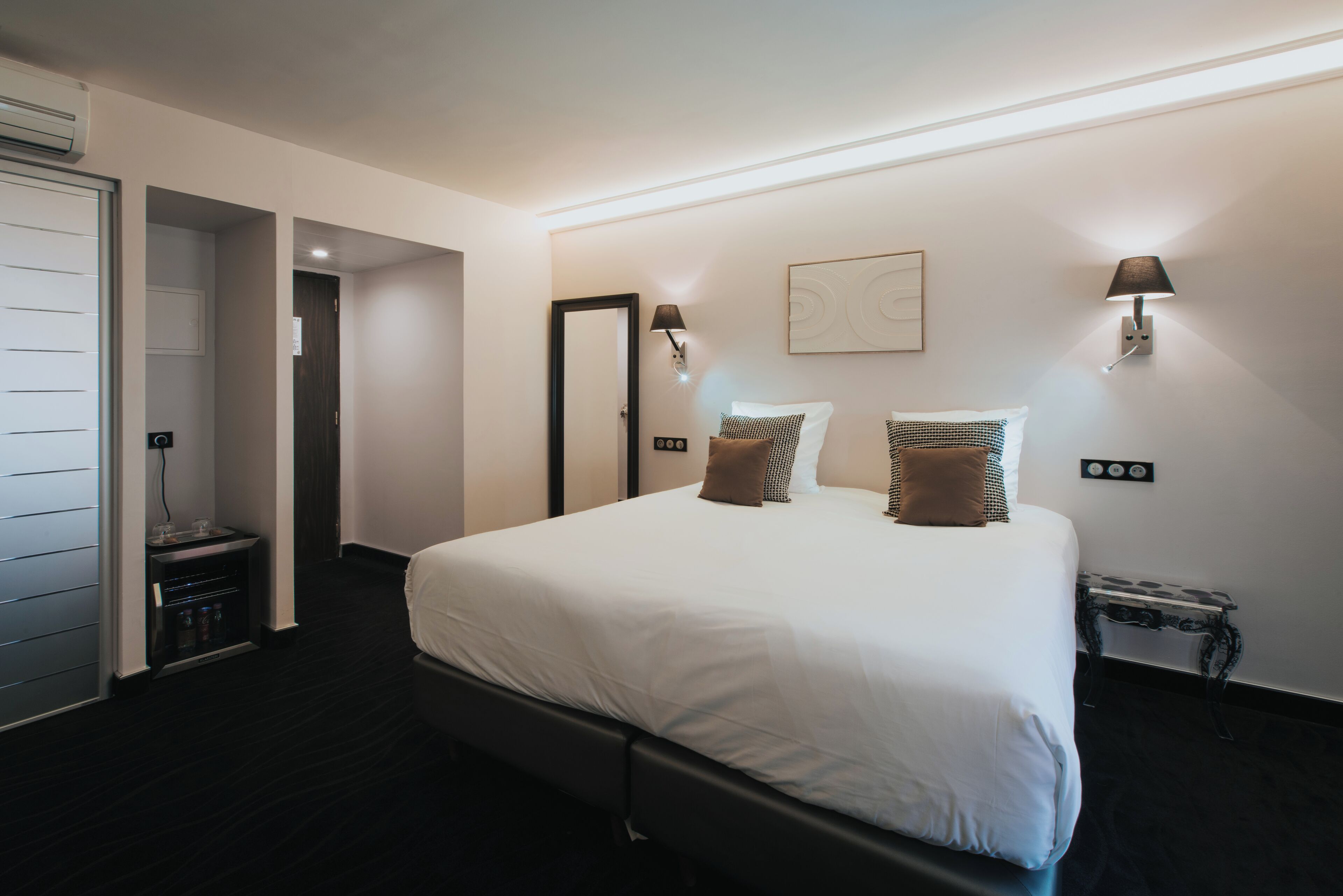 Une chambre d’hôtel moderne avec un grand lit, deux lampes fixées au mur, une petite table d’appoint et une porte donnant sur une salle de bain.
