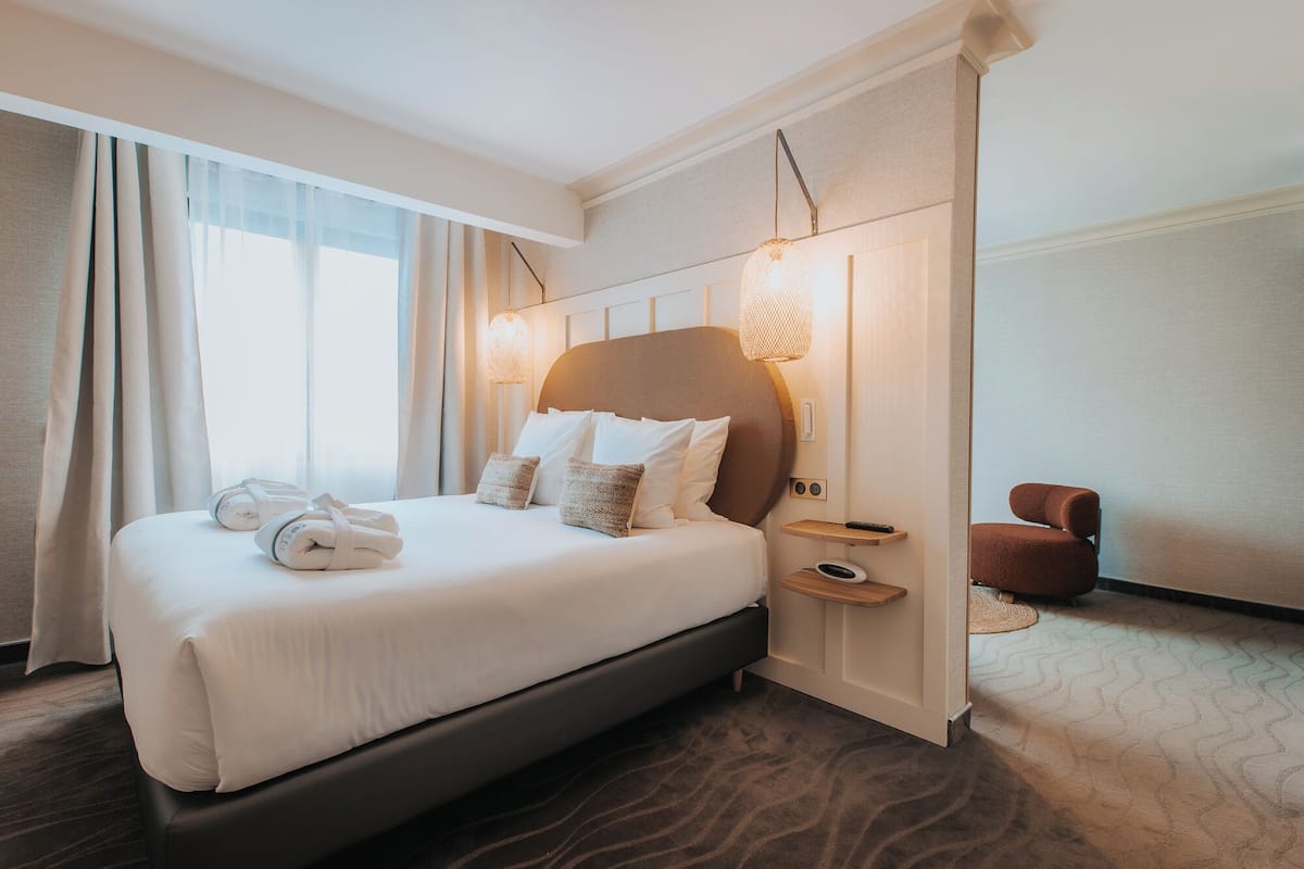 A modern hotel room with a large bed, a bedside table, a chair, and a view of the city.