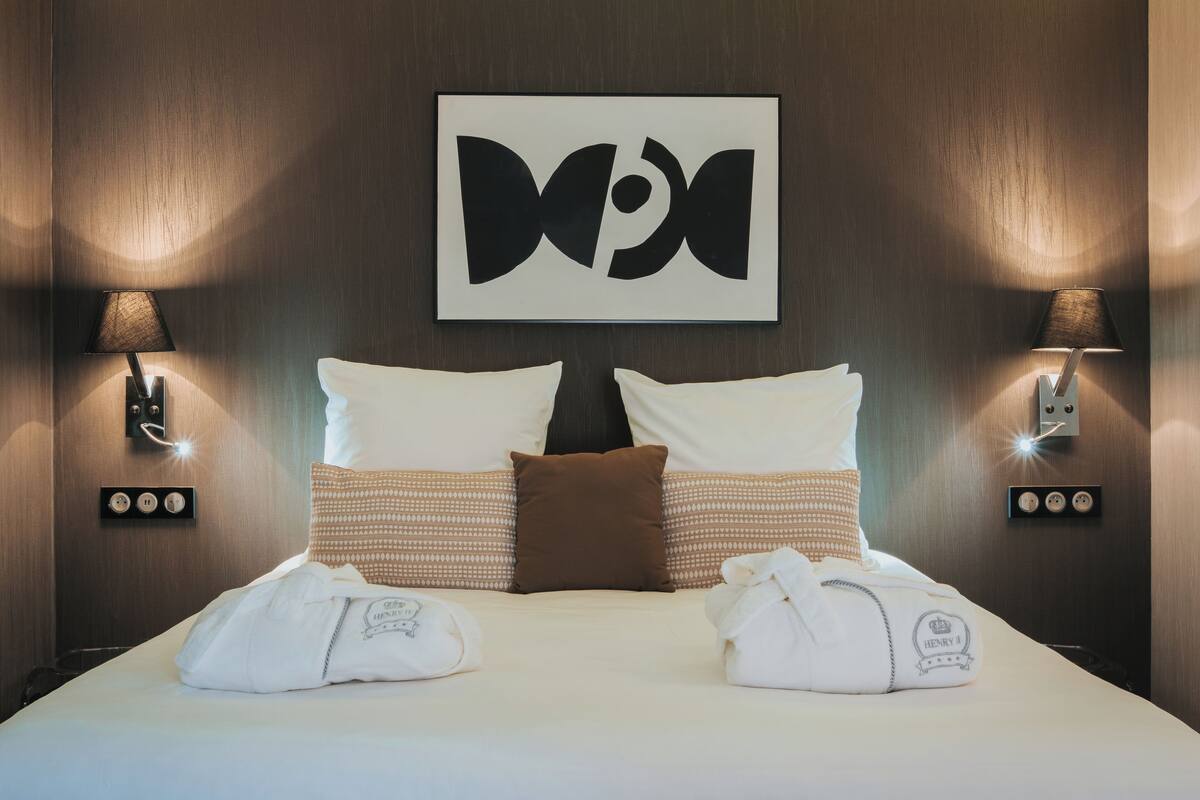 A hotel room with a bed, two pillows, two wall-mounted lamps, a wall-mounted artwork, and two towels with a monogram.