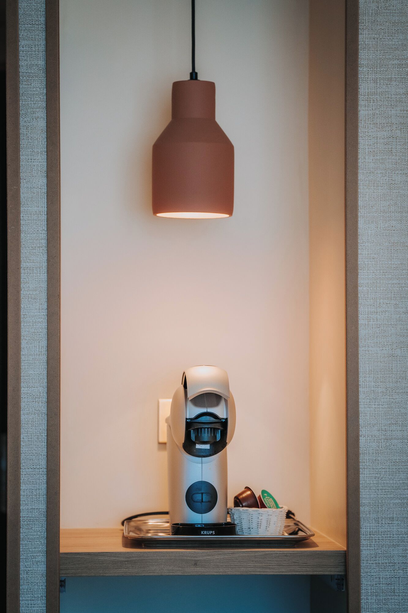 A coffee machine with a Krups brand label, a hanging pendant light, and a basket with items on a shelf.