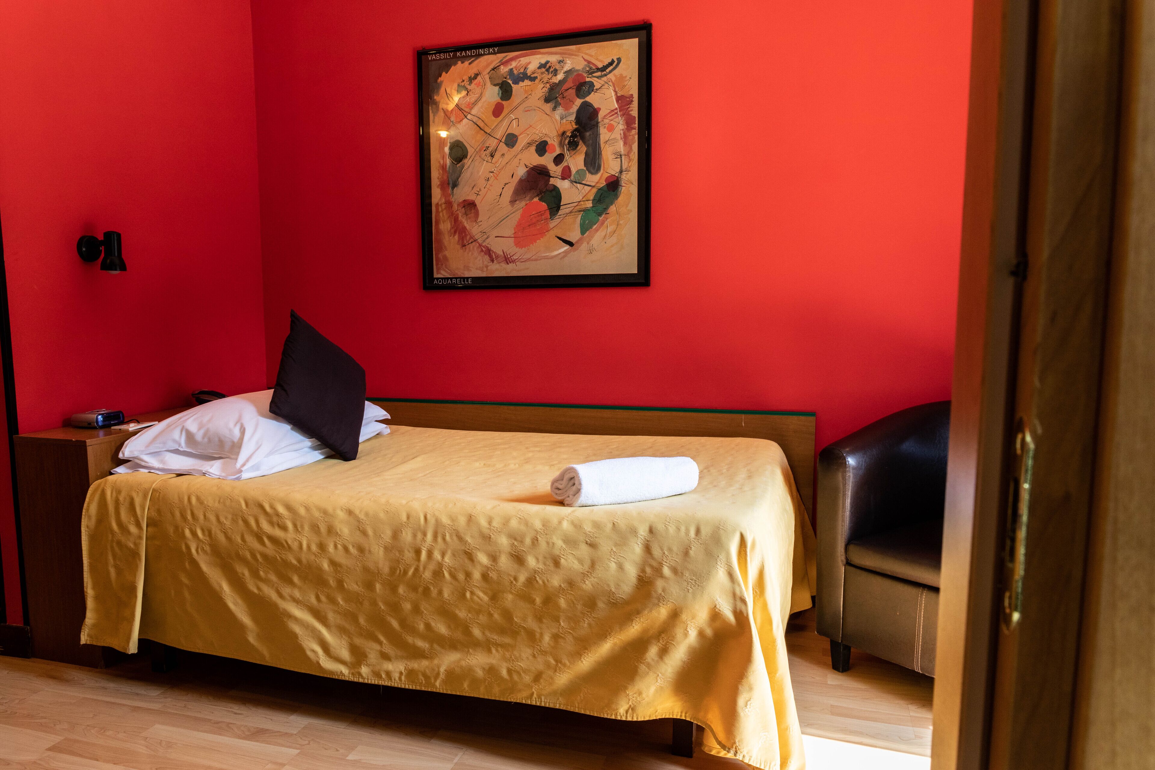A hotel room with a bed, a nightstand, a lamp, a painting on the wall, and a wooden door.