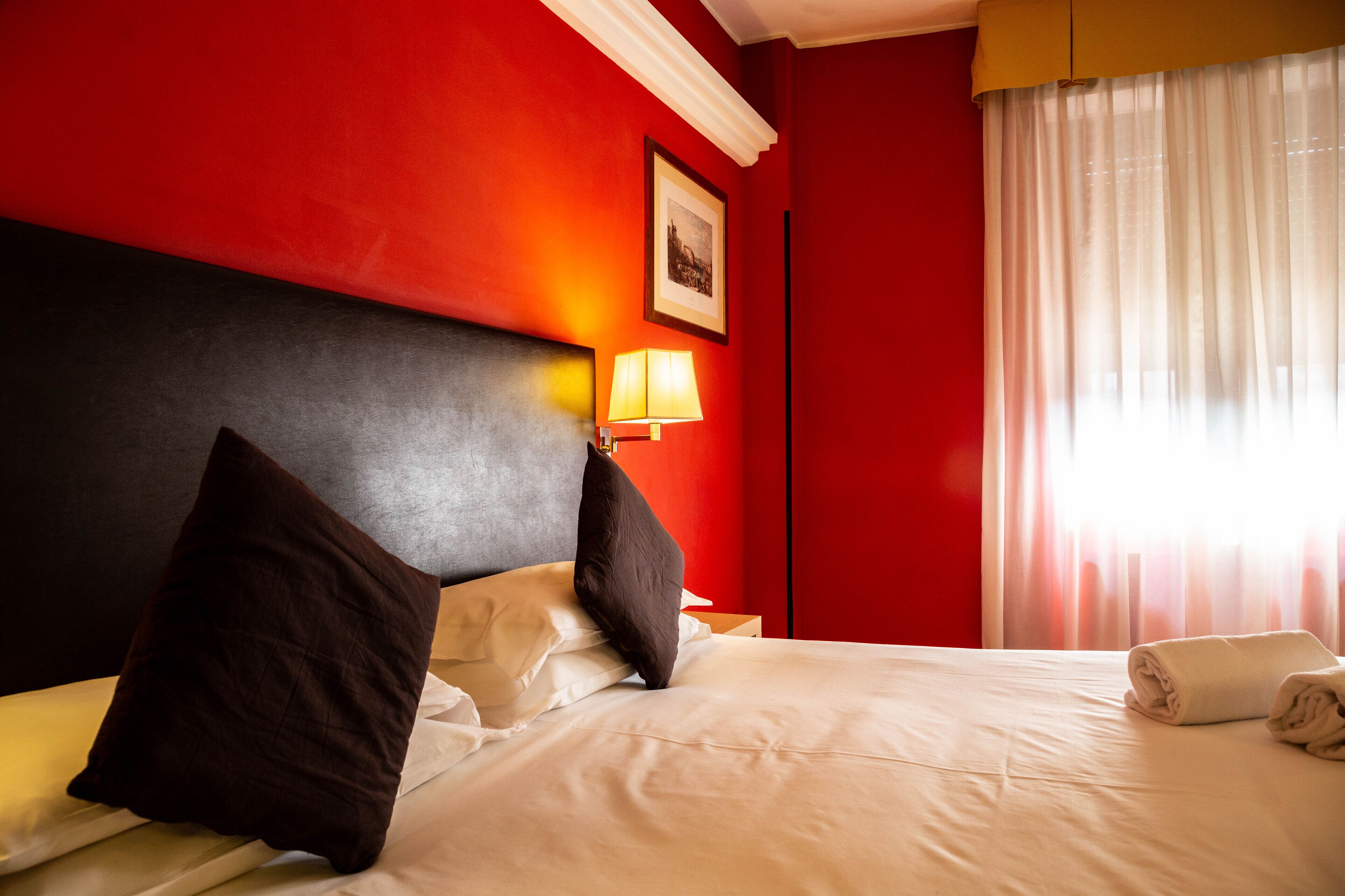 A hotel room with a red wall, a bed with white bedding, a black headboard, a lamp, and a window with curtains.