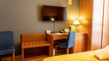 Una habitación de hotel con cama, un escritorio con silla, un televisor montado en la pared y una lámpara.