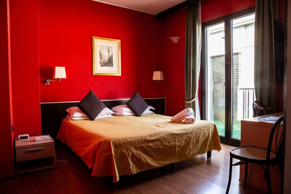 A hotel room with a bed, bedside lamps, a desk with a chair, and a window with curtains.