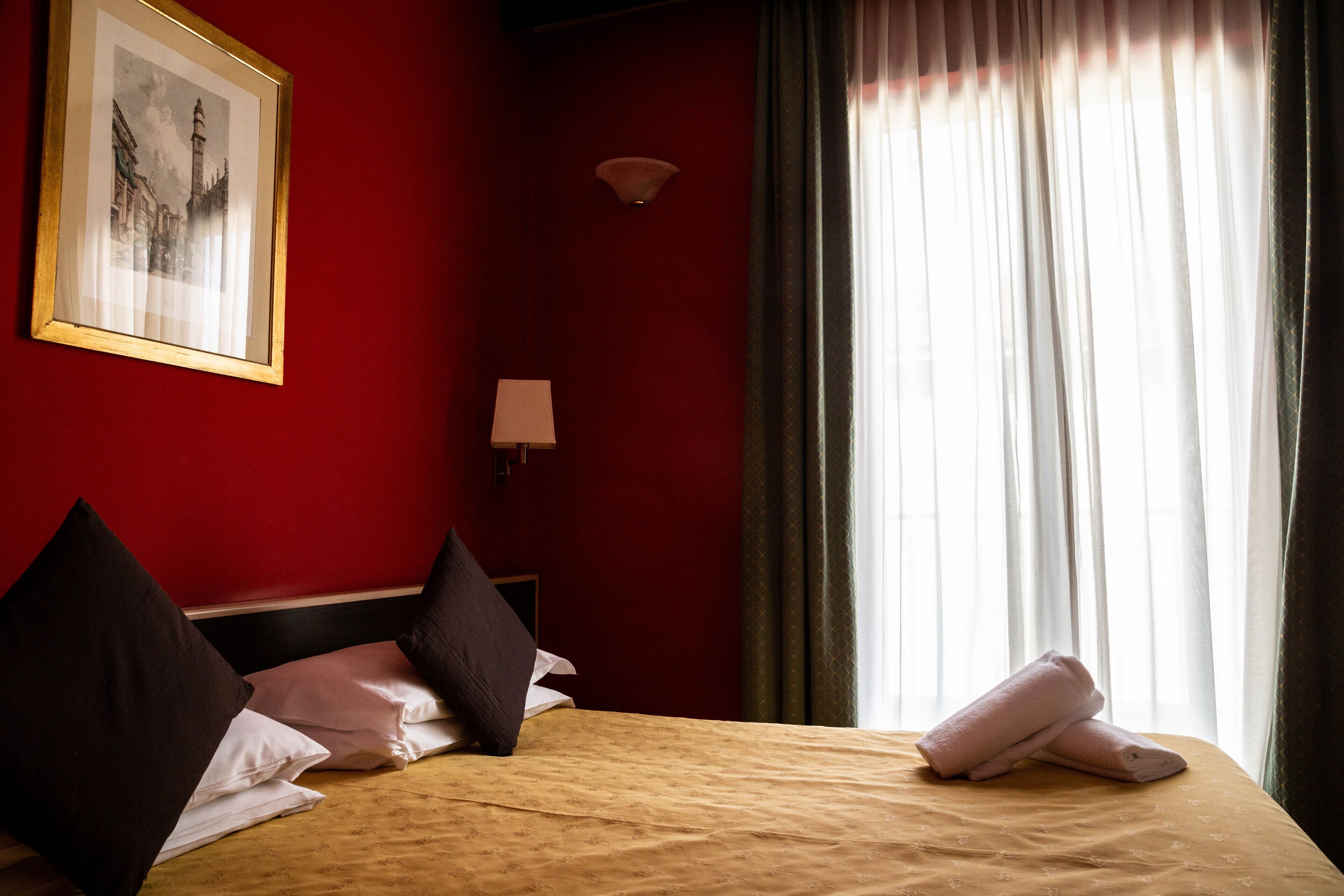 A hotel room with a bed, pillows, a framed picture on the wall, a bedside lamp, and a window with curtains.
