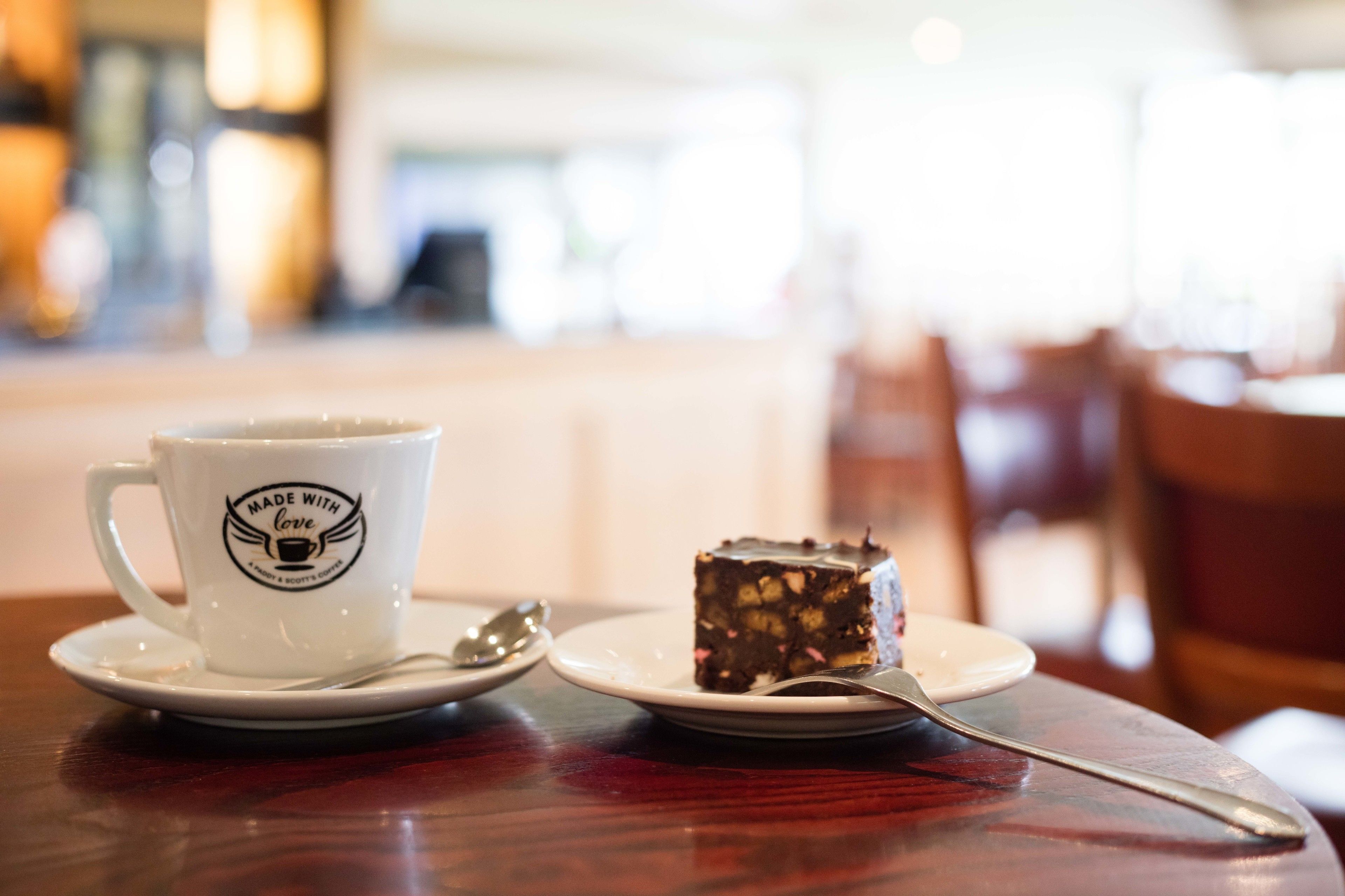 A white cup with a logo and text, a slice of cake on a plate, and a spoon on a wooden table.