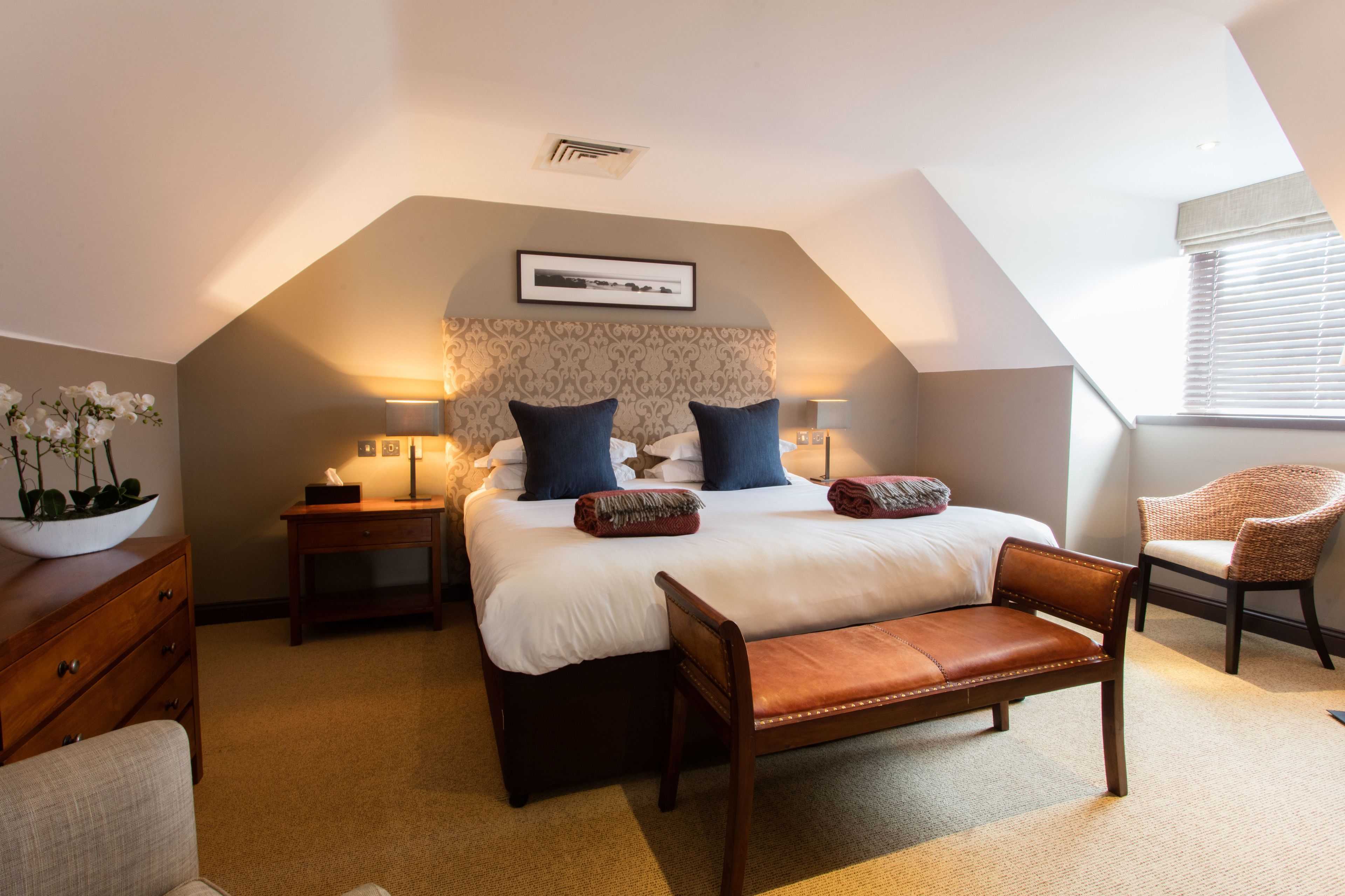 A bedroom with a large bed, a wooden bench, a chair, a nightstand, a lamp, and a framed picture on the wall.