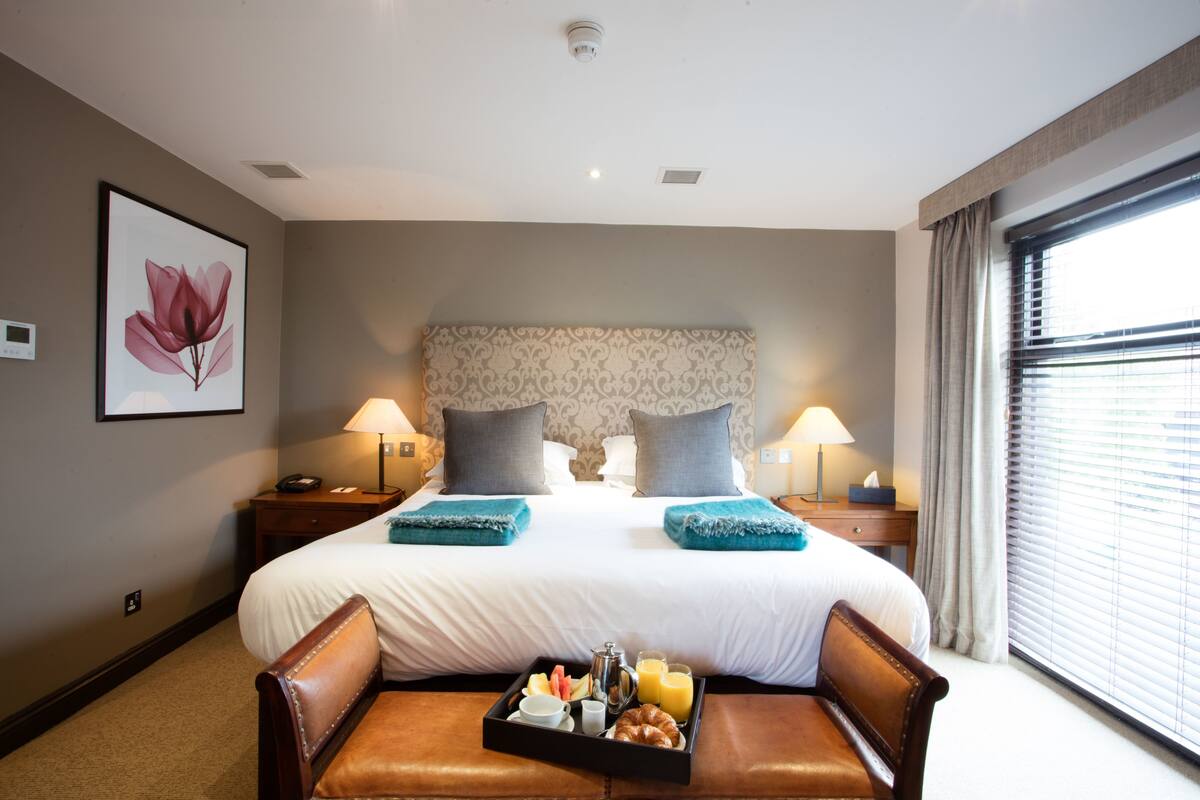 Une chambre d’hôtel avec un grand lit, deux tables de chevet avec des lampes, un plateau avec des articles pour le petit-déjeuner et une fenêtre avec des rideaux.