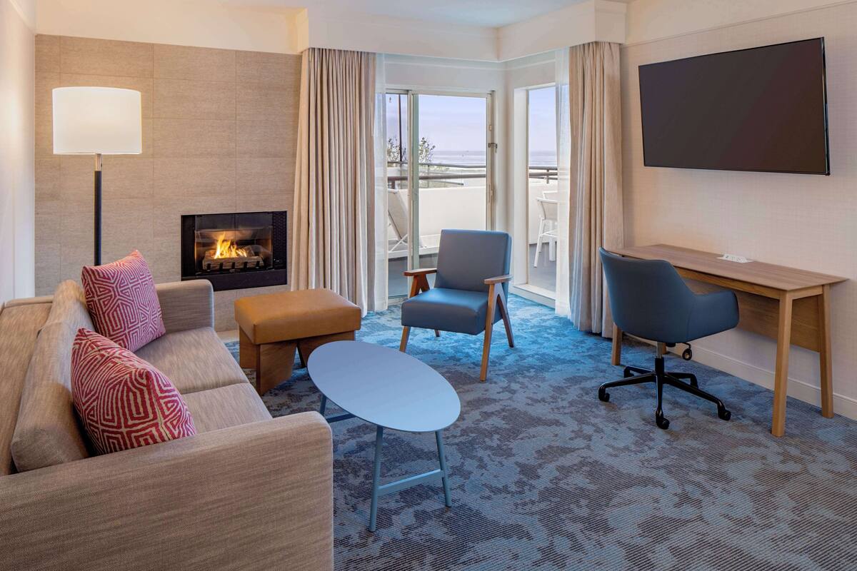 A hotel room with a sofa, ottoman, desk, chair, TV, fireplace, and a balcony with a view.