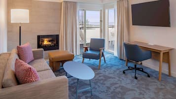 Habitación de hotel con sofá, reposapiés, escritorio, silla, televisor, chimenea y balcón con vistas.